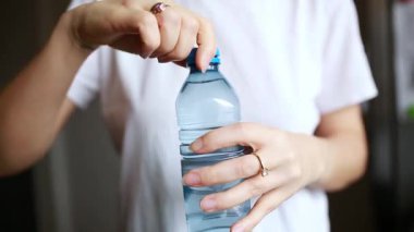 Arka planda bulanık çiçekler olan açık bir masadaki plastik şişeyi kapat. Genç bir kadının eli bağlı PET kapağını açar, günlük yaşamı, suyu ve çevre dostu kullanımı vurgular. Yüksek