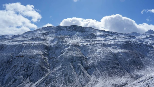 Karlı dağ sıraları, berrak mavi bir gökyüzüne karşı görkemli bir şekilde yükselir. Kabarık bulutlar, nefes kesici kış manzarasına derinlik katar.
