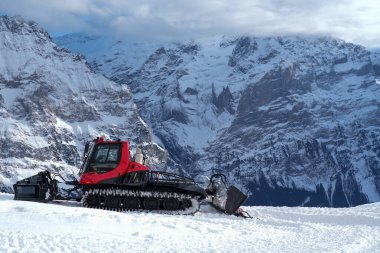 Kar temizleme aracı İsviçre Alpleri 'nde karlı bir yamaçta, yüksek dağ zirveleri ve berrak mavi gökyüzü çarpıcı bir kış manzarası yaratıyor.