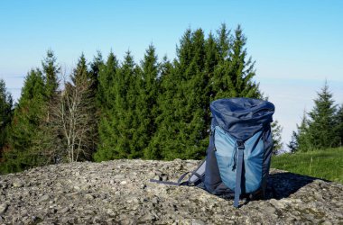 Mavi sırt çantası arka planda kozalaklı ağaçlarla kaplı kayalık bir yüzeye yerleştirilmiş açık mavi bir gökyüzü ve açık hava manzarası sergiliyor.