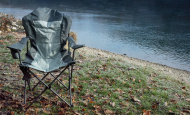 Sonbahar yaprakları ve sisli bir çevreyle çevrili sakin bir nehrin kenarındaki çimlere kurulmuş katlanır sandalye, sakin bir açık hava manzarası yaratıyor.