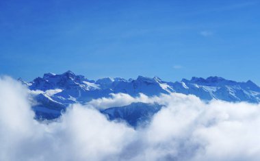 Kar kaplı dağ zirveleri bulutlardan oluşan bir denizin üzerinde berrak mavi bir gökyüzünün altında yükseliyor. Sıcaklığın tersine dönmesi dramatik bir alp panoraması yaratır, ideal bir seyahat ya da doğa arka planı olarak..