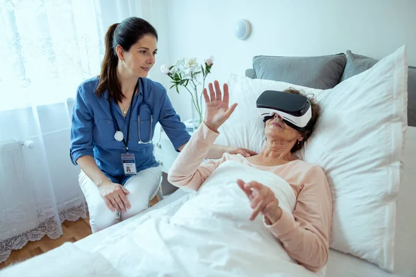 Kadın doktor, hastanede yatan yaşlı hastayı kontrol ediyor. Via VR Teknoloji ile terapi yapıyor. Arkadaş canlısı doktor kıdemli kadının sanal gerçeklik kulaklığı kullanarak terapi görmesine yardım ediyor..