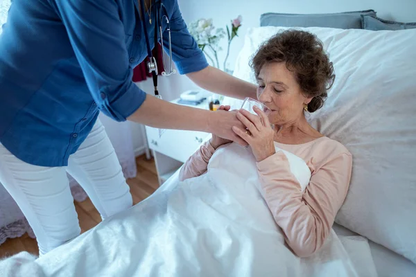 高齢患者の飲料水を助ける看護師 女性から水をもらうためにベッドに寝そべっている高齢女性介護者 ストック画像