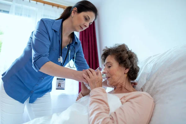 ホームキャリア高齢者の女性飲料水を支援します 看護師は 病気の高齢女性患者がベッドに横たわって水のグラスを飲むのを助ける ロイヤリティフリーのストック写真
