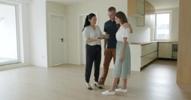 Yeni evde genç çiftlere dijital tablet gösteren bir emlakçı. Genç çift yeni evde dijital tabletle kadın emlakçıyla konuşuyor. Gerçek zamanlı, el kameralı, geniş açılı, 4K.