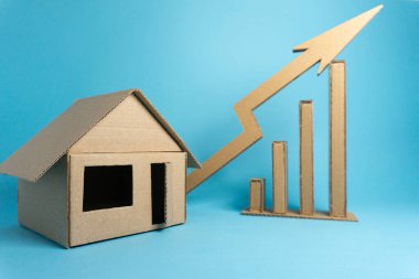 Bir evin mukavva modeli, kartondan yapılmış yükselen bir büyüme grafiğinin yanında yer alır. Sahne mavi bir arka plana göre ayarlandı. Bu kurulum gayrimenkul eğilimleri ve mali büyümeyi temsil etmektedir.