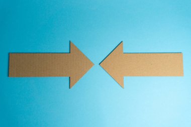 İki karton ok mavi bir yüzeyde karşı karşıya. Bu oklar kahverengi kartondan yapılmış ve açık bir alana yerleştirilmişler. Arka plan parlak ve basit..