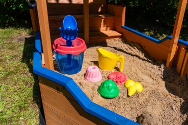 Parlak plastik oyuncaklarla dolu ahşap bir kum havuzu güneşin altında, pembe, kırmızı, yeşil ve sarı kalıpların yanında mavi bir kap ve kepçeyle duruyor. Canlı renkler ve neşeli şekiller açık hava eğlencesini ön plana çıkaran neşeli bir sahne yaratır.,