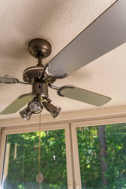 Modern tavan vantilatörünün dört pervaneli ve entegre spot lambasıyla yakın çekim görüntüsü. Vantilatör, geniş pencereli parlak bir odanın içindeki dokulu bir tavana monte edilerek doğal ışığın metalik detayları aydınlatmasına olanak sağlıyor. Görüntü