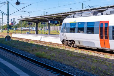 Modern bir bölgesel tren açık mavi gökyüzünün altındaki bir tren istasyonuna geliyor. Tepedeki elektrik hatları, metal yapılar ve çoklu raylar çağdaş demiryolu taşımacılığının detaylı bir görüntüsünü oluşturuyor. Arka planda başka bir tren görülebilir, EMP