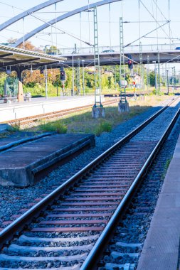 Görüntü, üstteki elektrik hatları ve çelik direklerle çerçevelenmiş bir istasyon bölgesinden geçen tren yollarını gösteriyor. Sol tarafta oturma, işaret ve sinyallere sahip bir platform, arka planda geniş bir köprü bulunmaktadır. Parlak gün ışığı.