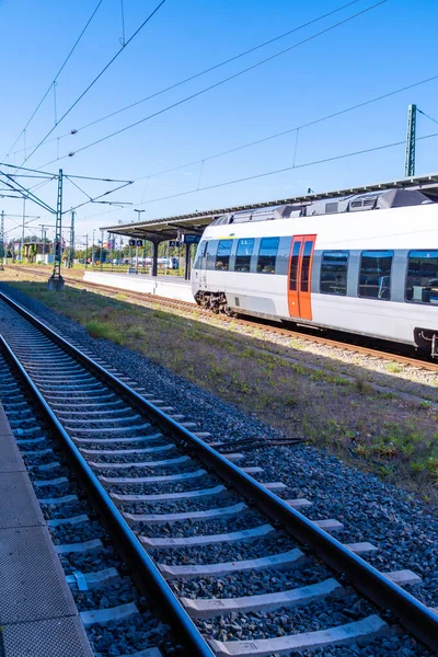Modern bir bölgesel tren, parlak mavi gökyüzünün altında bir tren istasyonunda duruyor. Tepedeki elektrik hatları ve çelik direkler sahneyi çerçevelerken, paralel raylar da mesafeye gidiyor. Görüntü temiz tren tasarımı, istasyon altyapısı,