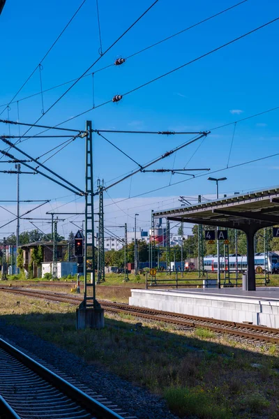 Birden fazla raydan oluşan demiryolu altyapısının detaylı bir görüntüsü, kablo hatları, sinyal ekipmanları ve bir istasyon platformu. Parlak mavi gökyüzü metal yapılarla tezat oluştururken, arka planda trenler ve binalar görülebilir. Th