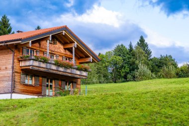 Yumuşak eğimli yeşil bir yamaçta büyüleyici bir ahşap dağ evi var. Evde kırsal ahşap yapı, kepenkli pencereler ve canlı kırmızı çiçeklerle süslenmiş balkonlar yer alıyor. Arkasında, bulutlu mavi gökyüzüne doğru yükselen sık ağaçlar,