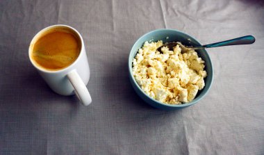 Tabakta taze süzme peynir, metal kaşık ve masada bir fincan kahve. Süt ürünleri ve sıcak içecekle sağlıklı kahvaltı kompozisyonu, doğal sabah yemeği natürmort yaşam, kırsal mutfak konsepti.