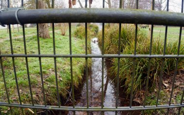 Çimenli nehir kıyıları arasında her iki tarafında ağaçlar olan küçük bir dere. Fotoğraf bir köprü korkuluğundan çekilerek doğal bir ön plan oluşturuluyor ve olay yerine derinlik ve perspektif ekleniyor. Sakin doğa manzarası.