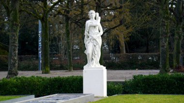 Düz bir park yolu, yolun kenarlarına yerleştirilmiş klasik heykellerle doludur. Taş figürler ağaçlar ve yeşillikler tarafından çerçevelenmiş resmi ve tarihi bir atmosfer yaratır. Yumuşak doğal ışık heykel detaylarını vurgular ve önderlik eder