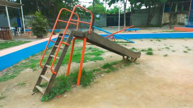 Parktaki ahşap kaydırağın üzerinde turuncu renkli bir merdiven var. Açık hava ortamında çocukların oynadığı mavi bir alanla çevrili yeşil çimenler var. Nostalji ve açık hava eğlencesi çağrıştırıyor. Aileler için mükemmel..