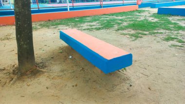 Oyun parkındaki uzun, renkli bir bank, mavi ve turuncu boyanmış, yerdeki kumla ve çevreleyen yeşillikle çelişiyor. Çevre, çocukların oynaması için neşeli ve canlı bir yer öneriyor..