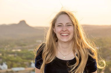 Neşeli bir kadın kameraya geniş bir gülümseme sergilerken, muhteşem bir dağ arkasındayken