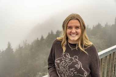 Sisli dağ manzarasının yanında rahat gezgin, sonbahar sabahı sıcacık giyinmiş neşeli bir kadının portresi