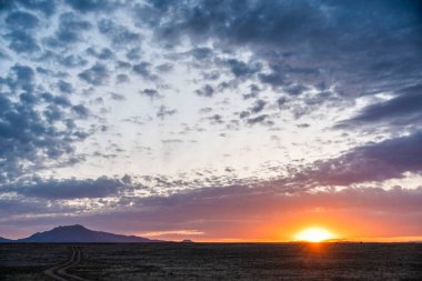 Güneş engin bir çöl manzarasının üzerinde zarifçe batarken, muhteşem dağların silueti arka planda güzelce yükseliyor.