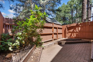 Dayanıklı ahşap bir çit güzel bir tuğla verandanın etrafını sararken, ağaçlar arka planda doğal gölgeler oluşturarak huzurlu ortamı güçlendiriyor.