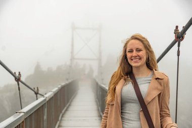 Şehir sisinin ortasında kendinden emin bir kadın gezgin, sise karşı ihtiyatlı bir güvenle köprüde sarışın bir kadın.