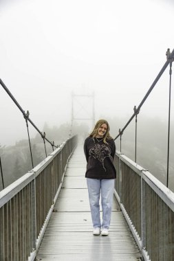 Şehir sisinin ortasında kendinden emin bir kadın gezgin, sise karşı ihtiyatlı bir güvenle köprüde sarışın bir kadın.