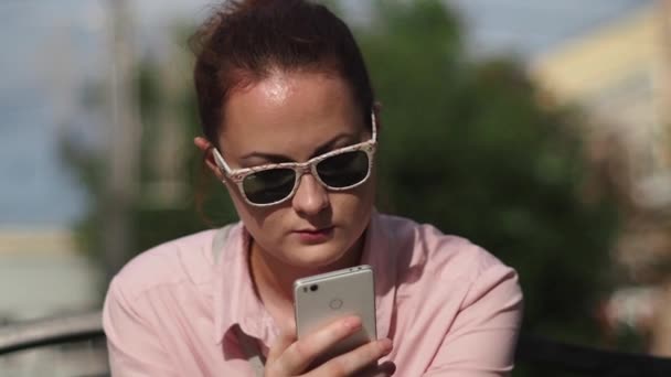 Молодая деловая леди в офисном стиле и в стильных солнцезащитных очках читает что-то с серьезным лицом на смартфоне в парке — стоковое видео