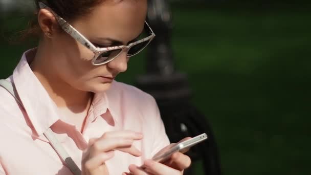 Mujer joven en una elegante blusa con cuello y gafas de sol elegantes escribiendo un mensaje en una pantalla táctil de un teléfono inteligente — Vídeos de Stock