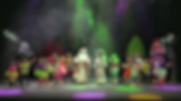 子供たちとサンタクロースは新年のコンサートで踊ります 雪のステージ上のサーチライトの多色ビーム 専用画像 — ストック動画