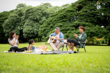 Mutlu Asyalı nesiller parkta pikniğin tadını çıkartıyor büyükbabam gitar çalıyor neşeli bir açık hava hayatı yaratıyor.