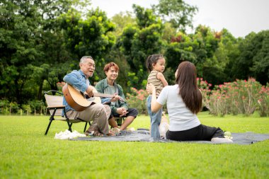Mutlu Asyalı nesiller parkta pikniğin tadını çıkartıyor büyükbabam gitar çalıyor neşeli bir açık hava hayatı yaratıyor.