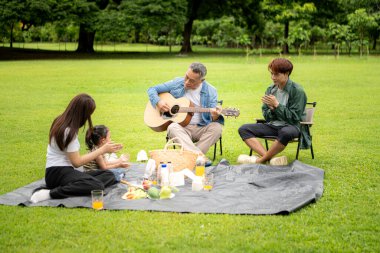Mutlu Asyalı nesiller parkta pikniğin tadını çıkartıyor büyükbabam gitar çalıyor neşeli bir açık hava hayatı yaratıyor.