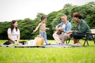 Mutlu Asyalı nesiller parkta pikniğin tadını çıkartıyor büyükbabam gitar çalıyor neşeli bir açık hava hayatı yaratıyor.