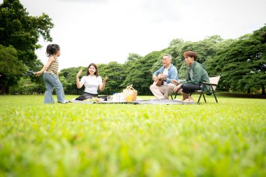 Mutlu Asyalı nesiller parkta pikniğin tadını çıkartıyor büyükbabam gitar çalıyor neşeli bir açık hava hayatı yaratıyor.