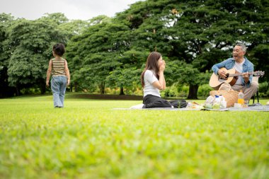 Mutlu Asyalı nesiller parkta pikniğin tadını çıkartıyor büyükbabam gitar çalıyor neşeli bir açık hava hayatı yaratıyor.