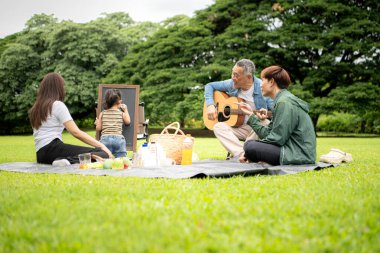 Mutlu Asyalı nesiller parkta pikniğin tadını çıkartıyor büyükbabam gitar çalıyor neşeli bir açık hava hayatı yaratıyor.