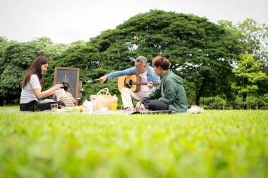 Mutlu Asyalı nesiller parkta pikniğin tadını çıkartıyor büyükbabam gitar çalıyor neşeli bir açık hava hayatı yaratıyor.