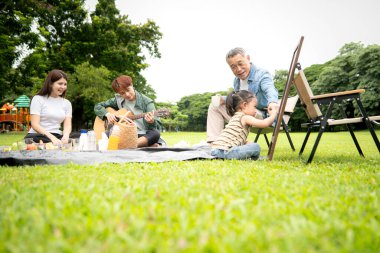 Mutlu Asyalı nesiller parkta pikniğin tadını çıkartıyor büyükbabam gitar çalıyor neşeli bir açık hava hayatı yaratıyor.