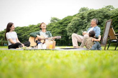 Mutlu Asyalı nesiller parkta pikniğin tadını çıkartıyor büyükbabam gitar çalıyor neşeli bir açık hava hayatı yaratıyor.