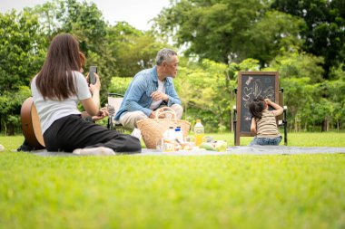 Mutlu Asyalı nesiller parkta pikniğin tadını çıkartıyor büyükbabam gitar çalıyor neşeli bir açık hava hayatı yaratıyor.