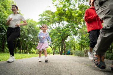 Asyalı bir aile parkta birlikte koşuyor. Ebeveynler, çocuklar ve büyükbabalar dışarıda egzersiz yapıyor, sağlıklı yaşam tarzını ve aile bağlarını destekliyor.