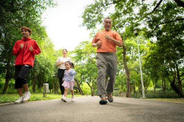 Asyalı bir aile parkta birlikte koşuyor. Ebeveynler, çocuklar ve büyükbabalar dışarıda egzersiz yapıyor, sağlıklı yaşam tarzını ve aile bağlarını destekliyor.