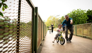 Büyükbaba küçük kıza parkta denge bisikleti sürmeyi öğretirken aile destek oluyor, sevgi, rehberlik ve aktif açık hava yaşam tarzı gösteriyor.