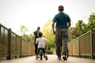 Büyükbaba küçük kıza parkta denge bisikleti sürmeyi öğretirken aile destek oluyor, sevgi, rehberlik ve aktif açık hava yaşam tarzı gösteriyor.