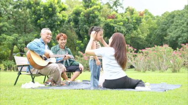 Mutlu Asyalı nesiller parkta pikniğin tadını çıkartıyor büyükbabam gitar çalıyor neşeli bir açık hava hayatı yaratıyor.