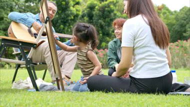 Mutlu Asyalı nesiller parkta pikniğin tadını çıkartıyor büyükbabam gitar çalıyor neşeli bir açık hava hayatı yaratıyor.